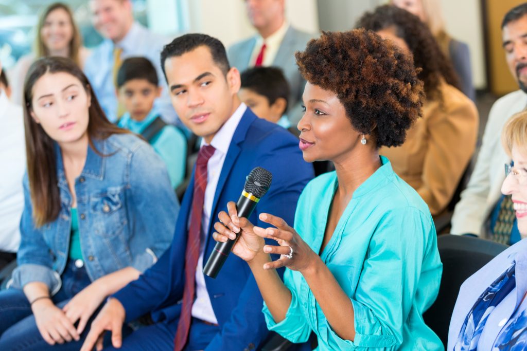 An African American woman sitting in an audience holds up a microphone as she asks a question to a speaker in front of her.