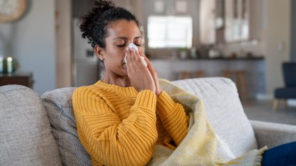 Portrait of young black woman sneezing in to tissue at home. Sick african woman wrapped in blanket sitting on sofa blowing her nose at home. Ill girl sneezing with runny nose in winter.